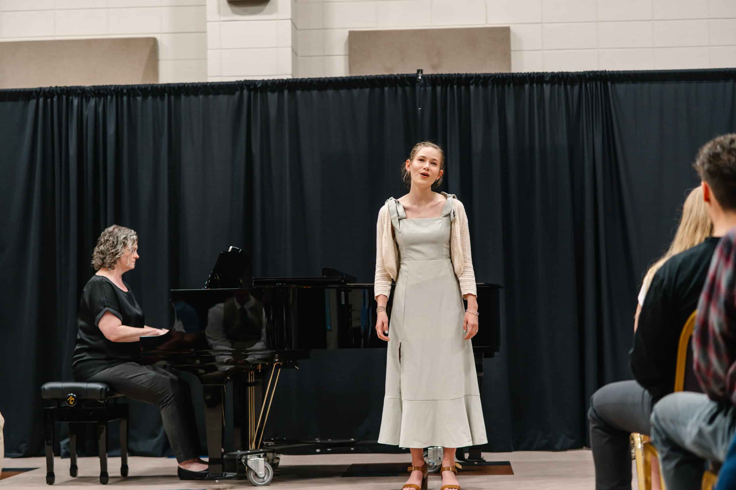 A student singing and a lady accompanying her with piano beside her.